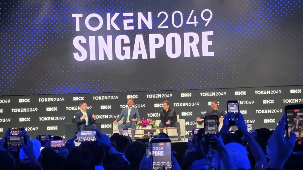 Sesión de World Liberty Financial «The Future of Finance, Built in America» en el escenario principal de OKX en TOKEN2049 Singapur 2026, con el público grabando con el móvil